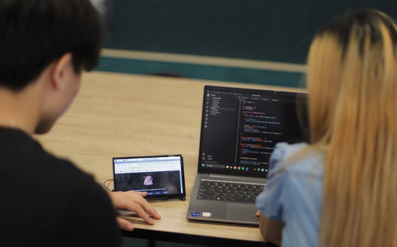 Two team members coding the Interactive Capybara, with Stella shown on a small screen beside the laptop
